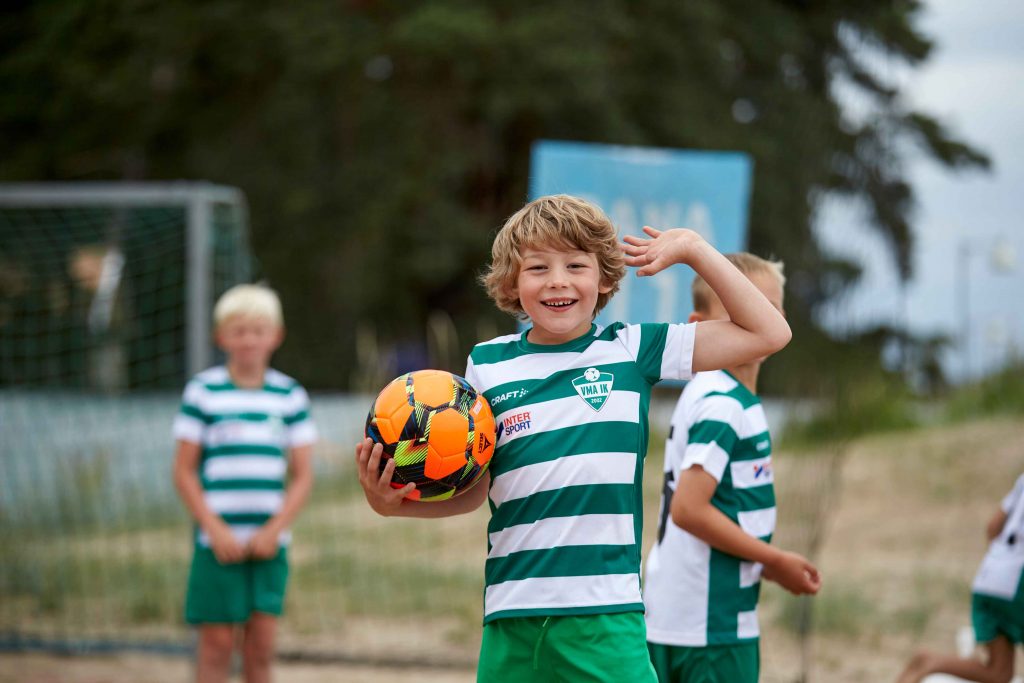 Glad ung kille poserar framför kameran med en fotboll i handen på Åhus Beachfotboll festival