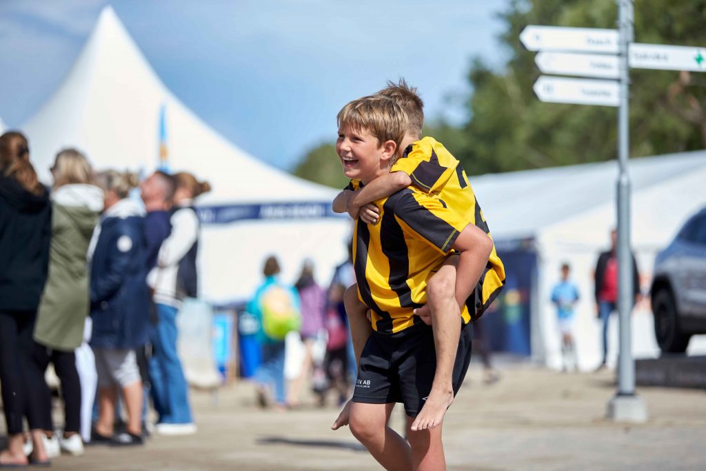 Glad pojke klädd i gult bärandes på sin vän på ryggen på Åhus Beachfotboll festival