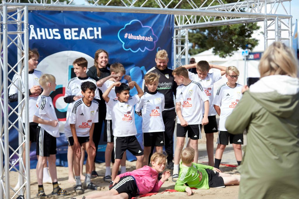 Helt fotbollslag med unga killar poserar framför Åhus Beachs tiktok-vägg på Åhus Beachfotboll festival