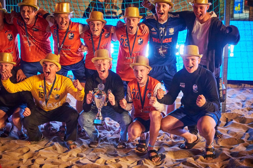Vinnare lag visar upp sin pokal framför ett handbollsmålvakt på Åhus Beach handbolls festival